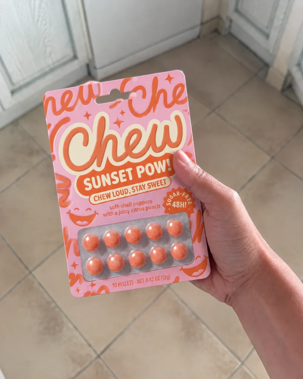 A hand holding a packet of Chew Sunset Pow gum over a tiled floor, taken on a phone.